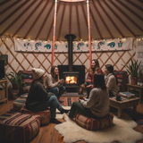 Group of people sitting around a fire in a yurt-like structure with wooden walls and decorative elements.