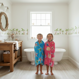 Two children standing in a bathroom with colorful towels, palm tree decals on the wall.