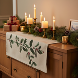Decorative table setting with candles, greenery, and a holly-patterned stenciled table runner on a wooden surface.