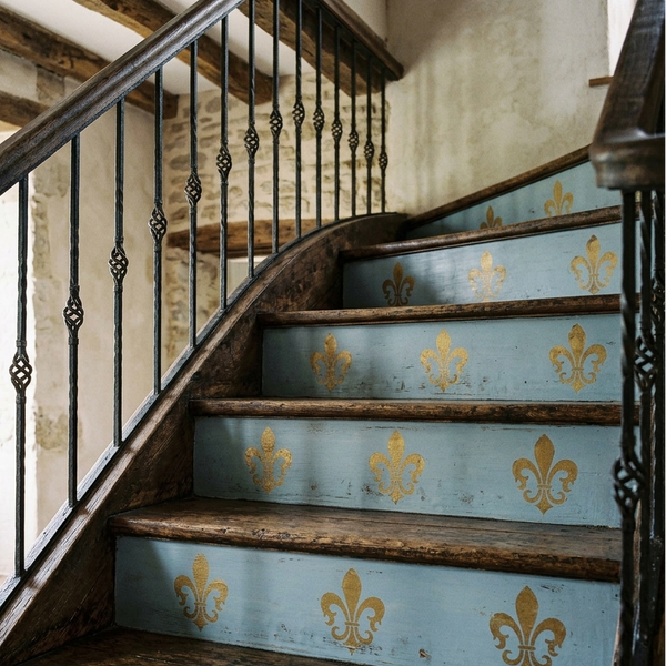 A rustic wooden staircase with light blue painted steps decorated with gold fleur-de-lis motifs, dark metal balusters, and a blue-and-white gingham runner at the bottom.