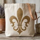 A natural canvas tote bag with a large gold fleur-de-lis printed on the front, featuring a tan leather shoulder strap, set on a rustic wooden surface with potted lavender in the background.
