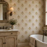 A styled bathroom interior featuring beige wallpaper with a repeating gold fleur-de-lis pattern, a vintage-style vanity with a mirror, wall sconce, and vase of flowers, plus a white freestanding tub with a towel draped over the edge.