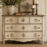 A distressed white and beige wooden dresser with gold fleur-de-lis accents on the drawer fronts, styled in a cozy room setting with a potted plant, candles, and framed art.
