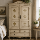 A tall distressed white armoire with gold fleur-de-lis accents on the doors, styled in a cozy bedroom next to a bed and a side table.