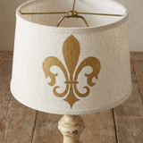 A table lamp shade made of textured off-white fabric with a large gold fleur-de-lis design, shown on a distressed cream-colored wooden lamp base over a rustic wooden tabletop.