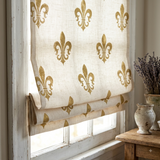 A beige linen Roman shade with a repeating gold fleur-de-lis pattern, partially drawn over a rustic white-framed window in a styled room setting.