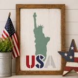 Framed artwork with Statue of Liberty silhouette and 'USA' text, American flag, and star decoration on a wooden surface.
