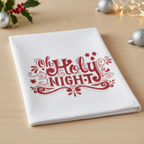 White towel with red 'Oh Holy Night' text on a wooden surface with Christmas decorations.