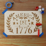 Stencil with 'America Est. 1776' on a wooden background with craft paints, roller and painter's tape