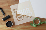 Stencil with 'you rock GRAD 2026' text on a wooden surface, with poster, paint and tape.