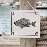 Decorative sign with fish design hanging on a wooden post by a body of water.