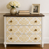 Decorative chest of drawers with gold pattern and gold knobs, featuring a vase with white flowers and books on top.