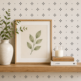 Framed artwork with leaves, vase with greenery, and candle on a shelf against a patterned wall.