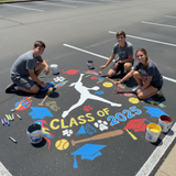 Three students painting 'Class of 2025' on a parking lot with various symbols.