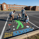 Four students painting a large design on the ground with 'Class of 2026 Seniors' text in a school parking lot.