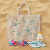 Beige tote bag with starfish design on sand, accompanied by a colorful bikini top, white flip-flops, and sunscreen bottle.