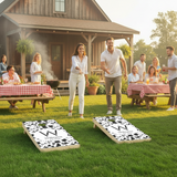 People playing stenciled cow spots cornhole in a backyard setting with a cabin in the background