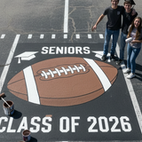 Three high school seniors standing on a large painted football in a parking lot with 'Seniors Class of 2026' text.