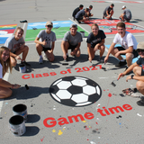 Group of people painting a large soccer ball and text on a paved surface, with 'Class of 2027' and 'Game time' visible.