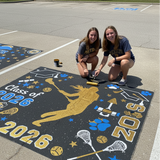 Two girls painting a large black mat with gold and blue designs, including 'Class of 2026' text, in a parking lot.