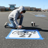 Person painting 'Class of 2027' on a parking lot with a school building in the background.
