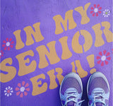 Person standing on pavement that has been stenciled with 'In My Senior Era!' text.